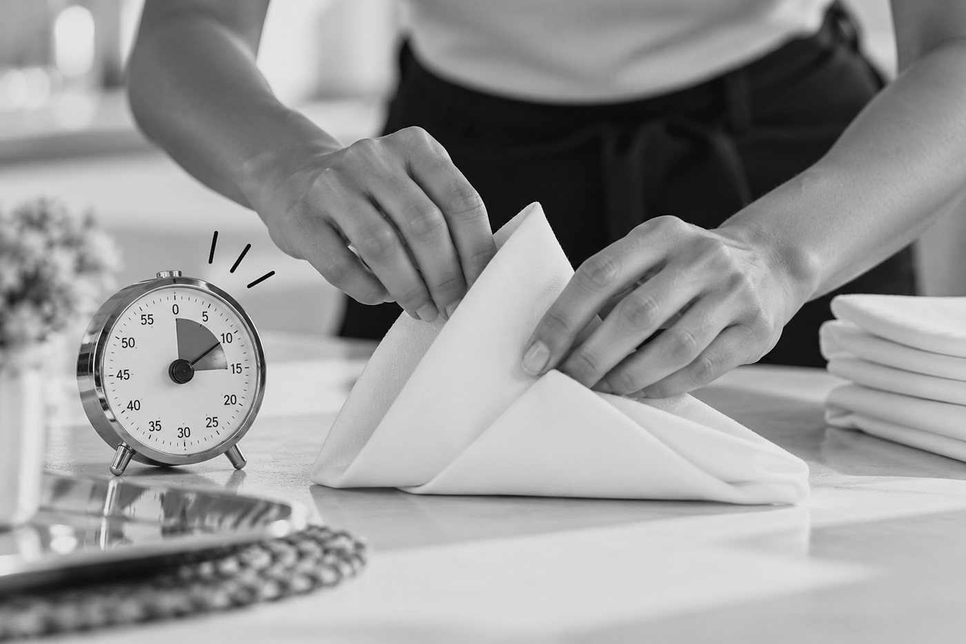 Close-up of hands folding a crisp white napkin beside a small kitchen timer and a stack of folded napkins