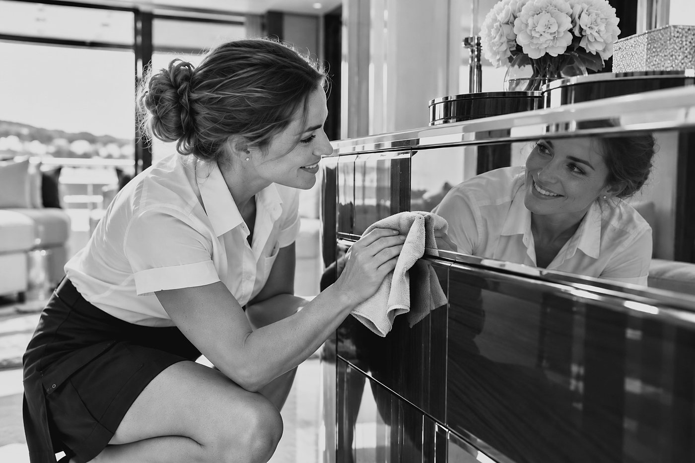 A stewardess crouching beside a high-gloss sideboard with a microfibre cloth, checking the surface against the light as her smiling reflection looks back