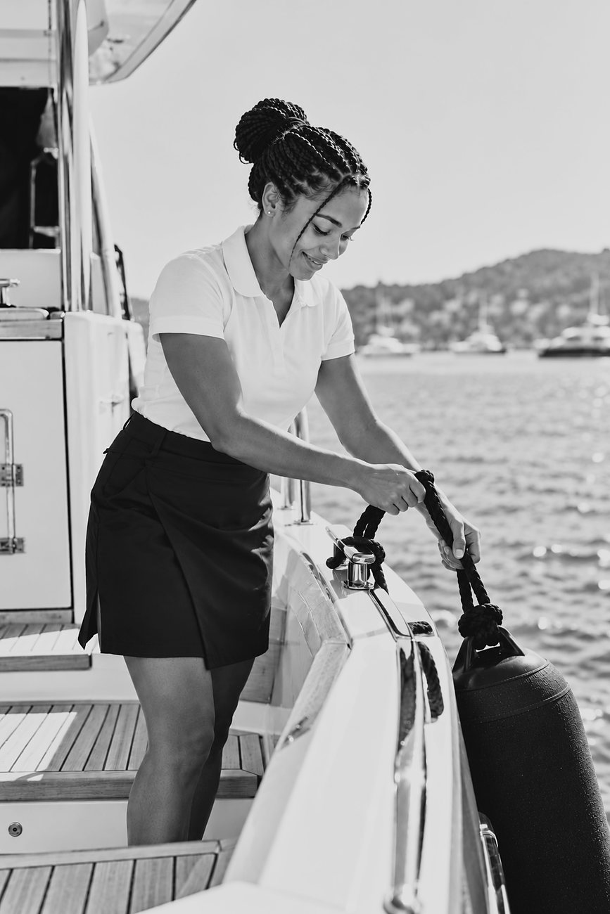 A smiling stewardess in a white polo shirt and navy skort tying off a fender line on a cleat at the side deck of a yacht, with the sea and hills behind her